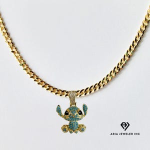 May include: Gold-colored chain necklace with a pendant featuring a cartoon character with blue and black accents. The pendant is encrusted with small, light blue stones. The necklace is displayed on a white background.
