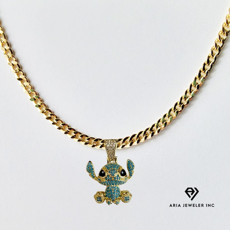 May include: Gold-colored chain necklace with a pendant featuring a cartoon character with blue and black accents. The pendant is encrusted with small, light blue stones. The necklace is displayed on a white background.
