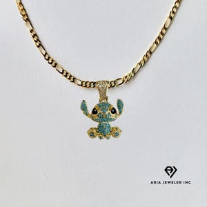 May include: A gold-colored chain necklace with a pendant featuring a cartoon character. The pendant is a blue and gold-colored character with black eyes and is encrusted with small, sparkling stones. The necklace is on a white background.