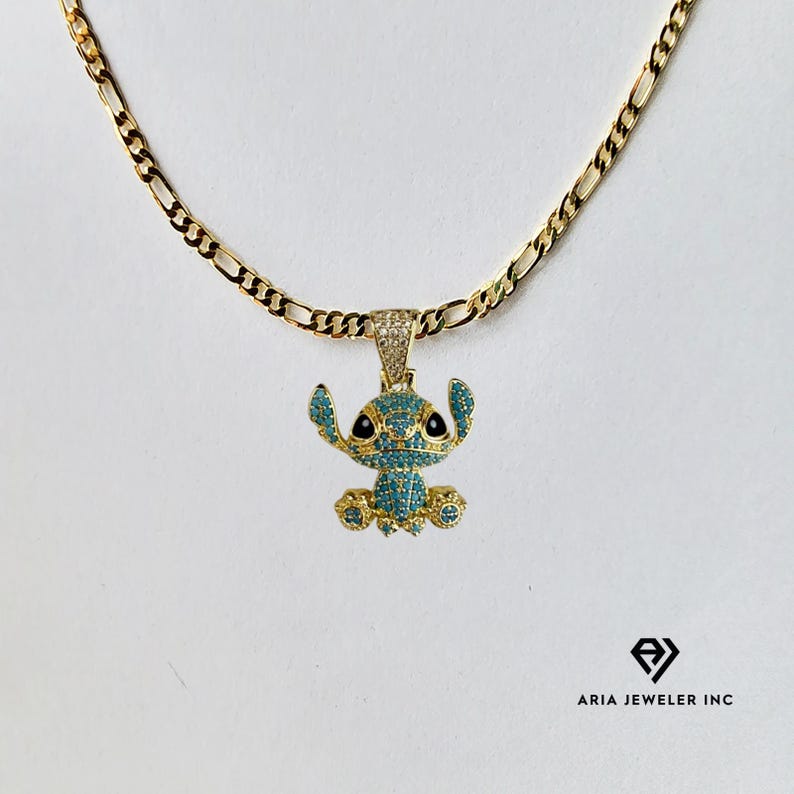 May include: A gold-colored chain necklace with a pendant featuring a cartoon character. The pendant is a blue and gold-colored character with black eyes and is encrusted with small, sparkling stones. The necklace is on a white background.