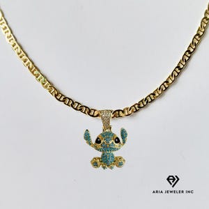 May include: A gold-colored chain necklace with a pendant featuring a blue and gold character design. The pendant is encrusted with small, sparkling stones. The necklace is displayed on a white background.