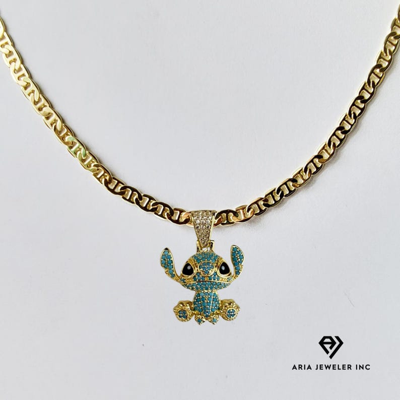 May include: A gold-colored chain necklace with a pendant featuring a blue and gold character design. The pendant is encrusted with small, sparkling stones. The necklace is displayed on a white background.