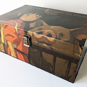 May include: A wooden box with a hinged lid and a metal clasp. The box is decorated with a colourful illustration of a young woman with long dark hair and a red dress, standing next to a large pig. The text "Chihiros Reise ins Zauberland" is printed on the top of the box.