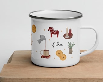 May include: White enamel mug with a silver rim, featuring illustrations of a coffee pot, a Dala horse, candles, and pastries. The word "fika" is written in black script. The mug is on a wooden cutting board.