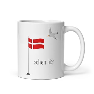 Könnte beinhalten: Weiße Keramiktasse mit einer rot-weißen dänischen Flagge und einer Möwe, die darüber fliegt. Der Text "schön hier" ist auf die Tasse gedruckt.