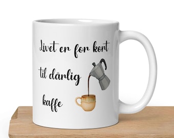 May include: White ceramic coffee mug with the text "Livet er for kort til dårlig kaffe" and a graphic of a coffee pot pouring into a mug. The mug has a white handle and is sitting on a wooden surface.