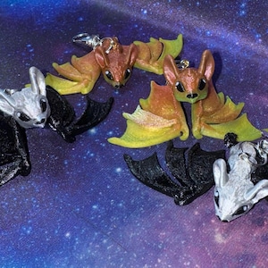 May include: Three pairs of bat earrings. One pair is black, one is orange and yellow, and one is silver and black.