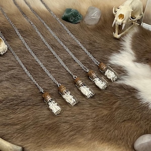 May include: Five silver chain necklaces, each with a small glass vial pendant filled with white bone fragments. The vials have cork stoppers. The necklaces are displayed on a brown and white fur-like surface, with animal skulls and stones.