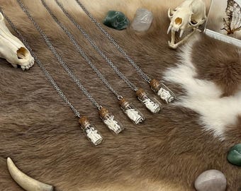 Könnte beinhalten: Fünf Halsketten mit silberner Kette, jeweils mit einem kleinen Glasfläschchen-Anhänger, gefüllt mit weißen Knochenfragmenten. Die Fläschchen haben Korken. Die Halsketten werden auf einer braun-weißen, fellartigen Oberfläche mit Tierschädeln und Steinen präsentiert.
