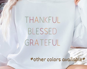 Peut inclure: Sweat-shirt blanc avec les mots "THANKFUL BLESSED GRATEFUL" en différentes couleurs. Le texte est en format empilé. Le bas du sweat-shirt a le texte "*other colors available*" en marron.