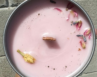 May include: Pink scented candle with dried flowers and a wooden wick. The candle is in a silver tin.