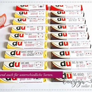 Puede incluir: Una colecci&oacute;n de barras de chocolate con envoltorios personalizados que presentan varias frases en alem&aacute;n e ilustraciones de animales lindos. Los envoltorios son blancos con texto rojo y negro, y las barras de chocolate tienen embalaje amarillo y marr&oacute;n. Se ve una mariposa de papel.
