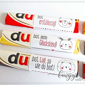 Puede incluir: Tres barras de chocolate con envoltorios personalizados. Los envoltorios presentan la palabra "du" en rojo, y cada uno tiene un mensaje y un animal de dibujos animados diferente: un oso, un conejito y un le&oacute;n. El texto de los envoltorios incluye "erstklassig!", "Gl&uuml;ckskind!" y "bist toll, so wie du bist!"