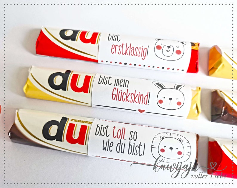 Puede incluir: Tres barras de chocolate con envoltorios personalizados. Los envoltorios presentan la palabra "du" en rojo, y cada uno tiene un mensaje y un animal de dibujos animados diferente: un oso, un conejito y un le&oacute;n. El texto de los envoltorios incluye "erstklassig!", "Gl&uuml;ckskind!" y "bist toll, so wie du bist!"