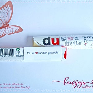 Puede incluir: Una tarjeta blanca con una mariposa coral y dos barras de chocolate. Una barra tiene la palabra "du" en rojo. La otra barra tiene una ilustraci&oacute;n de cebra y el texto "bist mehr als deine Noten!". La tarjeta tambi&eacute;n tiene texto en alem&aacute;n.