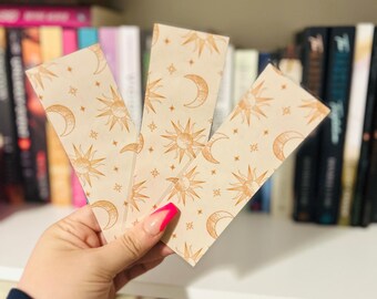 May include: Three celestial-themed bookmarks with a repeating pattern of suns, moons, and stars in a warm, golden colour. The bookmarks are held in front of a bookshelf filled with books.