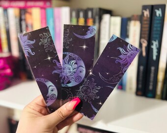May include: Three celestial-themed bookmarks with sun and moon designs in purple, blue, and white. The bookmarks are held in front of a bookshelf filled with books.