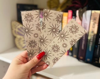 May include: Three beige bookmarks with a repeating sun and star pattern are held in front of a bookshelf. The sun design has a smiling face. The bookmarks are rectangular and have a glossy finish. The background includes a white shelf and a collection of books.