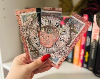 May include: A hand holds several vintage-style bookmarks. Each bookmark features a clock face design with Roman numerals, floral accents, and a distressed, aged appearance. The colour palette includes soft pinks, creams, and browns.