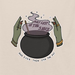 May include: Beige t-shirt featuring a graphic of a black cauldron with purple smoke and the text "GET LOST IN THE SAUCE". Green hands with gold jewelry surround the cauldron. The text "AND EVEN THEN STIR THE POT" is below the cauldron.