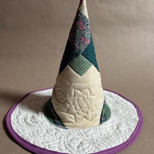 May include: A conical hat constructed from quilted fabric patches in shades of cream, dark green, and floral patterns. The wide brim is white with intricate quilted swirl stitching and finished with a solid purple fabric border trim.