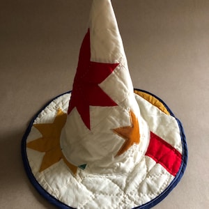 Puede incluir: Un sombrero de mago blanco acolchado con detalles de estrellas rojas y naranjas. El sombrero tiene un borde azul y adornos amarillos y rojos. El sombrero está hecho de tela y tiene un patrón acolchado.