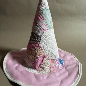 May include: A conical hat constructed from various quilted fabric patches in shades of white, pink, and floral patterns. The wide, circular brim is solid light pink with white edge binding, and the entire piece features visible quilted stitching.