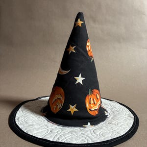 May include: A pointed witch hat featuring a black fabric cone printed with orange jack-o'-lanterns, yellow stars, and a crescent moon. The wide, flat brim is white with intricate, swirling quilted stitching and a solid black fabric edge trim.