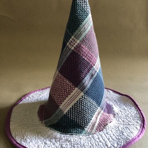 May include: A handmade witch hat with a plaid pattern in shades of blue, purple, and green. The hat has a wide, quilted brim with a white fabric and a purple trim. The hat is designed for costume or decorative purposes.