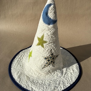 May include: A white quilted cone-shaped hat featuring a blue crescent moon appliqué and several stars in lime green and a patterned fabric. The hat has a wide brim with a dark blue fabric trim and intricate swirling stitch patterns throughout.