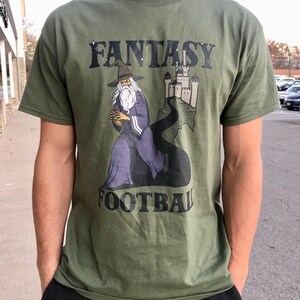 May include: Olive green t-shirt featuring the words "FANTASY FOOTBALL" above and below an illustration of a wizard holding a football. The wizard is wearing a hat and robe, with a castle in the background.