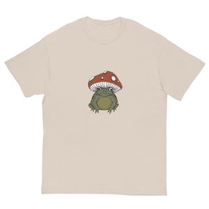 May include: A beige t-shirt with a cartoon illustration of a green frog wearing a red and white mushroom cap.