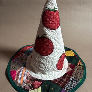 May include: A conical fabric hat featuring a white quilted crown decorated with three red tomato appliqués. The wide brim is constructed from a patchwork of various patterned fabric squares in shades of brown, green, and yellow, finished with a green trim.