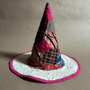 May include: A conical hat constructed from a patchwork of various fabric scraps in shades of hot pink, deep red, brown, and blue. The wide, circular brim is made of white quilted fabric with a hot pink border trim along the outer edge.