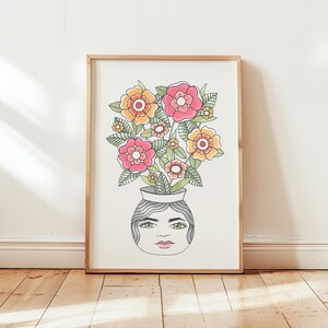 Puede incluir: Una impresión enmarcada de una ilustración colorida de un jarrón con una cara y flores. El jarrón es blanco con un contorno negro y las flores son rosas, amarillas y verdes.