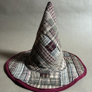May include: A conical hat constructed from quilted fabric patches in various plaid patterns, including shades of burgundy, olive green, cream, and navy blue. The wide, circular brim is finished with a solid burgundy fabric binding along the edge.
