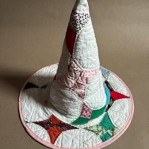May include: A conical hat constructed from quilted fabric patches. The design features a white base with various floral and geometric patterns in red, pink, green, and blue. The wide brim is finished with a solid light pink fabric binding edge.