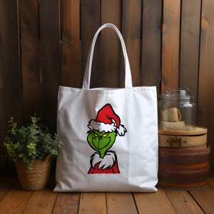 May include: White canvas tote bag with a green Grinch character wearing a red Santa hat. The Grinch is looking to the right with a mischievous expression.