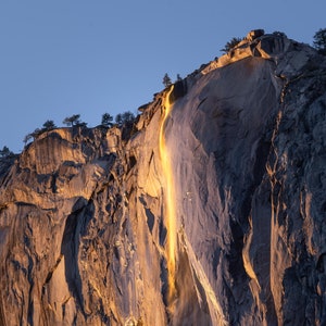 May include: A golden waterfall cascades down a sheer cliff face, illuminated by the setting sun. The rock face is a mix of gray and brown, with a rough texture. The waterfall is a thin stream of water, but it is brightly lit by the sun, making it appear like a golden ribbon.