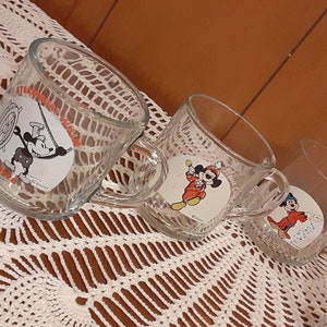 Puede incluir: Cuatro tazas de vidrio transparente con dise&ntilde;os de personajes de dibujos animados. Las tazas representan a Mickey Mouse, Donald Duck y Goofy.