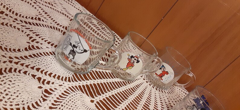 Puede incluir: Cuatro tazas de vidrio transparente con dise&ntilde;os de personajes de dibujos animados. Las tazas representan a Mickey Mouse, Donald Duck y Goofy.