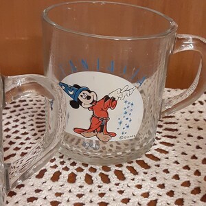 Puede incluir: Una taza de vidrio transparente con una ilustraci&oacute;n azul y blanca de Mickey Mouse como mago. La taza tiene el texto "Fantasia" y "1940" en ella. La taza est&aacute; sentada sobre un encaje de ganchillo blanco.