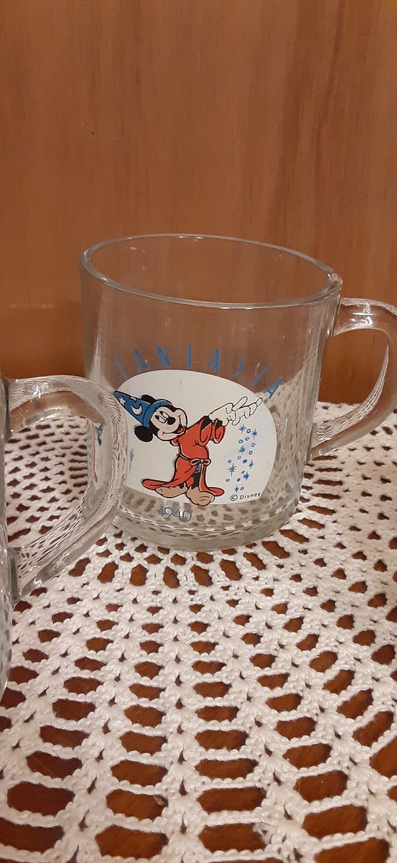 Puede incluir: Una taza de vidrio transparente con una ilustraci&oacute;n azul y blanca de Mickey Mouse como mago. La taza tiene el texto "Fantasia" y "1940" en ella. La taza est&aacute; sentada sobre un encaje de ganchillo blanco.