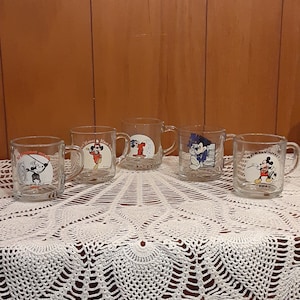 Puede incluir: Cinco tazas de vidrio transparente con dise&ntilde;os de Mickey Mouse. Cada taza presenta una imagen diferente de Mickey Mouse en una pose diferente. Las tazas est&aacute;n dispuestas en una fila sobre un mantel de crochet blanco.