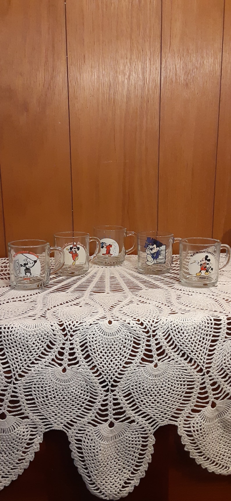 Puede incluir: Cinco tazas de vidrio transparente con dise&ntilde;os de Mickey Mouse. Cada taza presenta una imagen diferente de Mickey Mouse en una pose diferente. Las tazas est&aacute;n dispuestas en una fila sobre un mantel de crochet blanco.