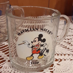 Puede incluir: Una taza de vidrio transparente con asa presenta un c&iacute;rculo blanco con una imagen de dibujos animados de Mickey Mouse vestido de mago. El texto "Magician Mickey" est&aacute; impreso sobre la imagen y "1937" est&aacute; impreso debajo de la imagen. El texto "&copy; Disney" est&aacute; impreso en la esquina inferior derecha del c&iacute;rculo.