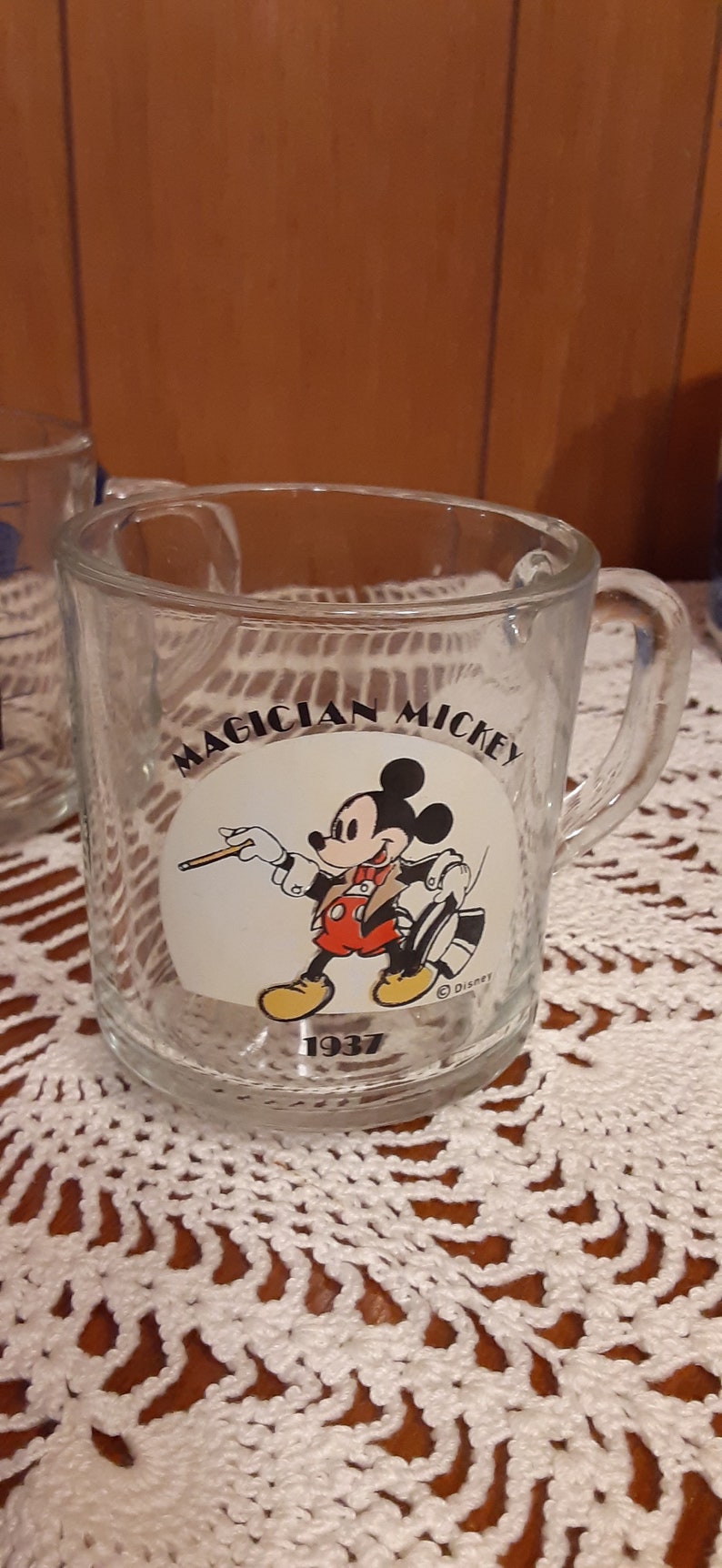 Puede incluir: Una taza de vidrio transparente con asa presenta un c&iacute;rculo blanco con una imagen de dibujos animados de Mickey Mouse vestido de mago. El texto "Magician Mickey" est&aacute; impreso sobre la imagen y "1937" est&aacute; impreso debajo de la imagen. El texto "&copy; Disney" est&aacute; impreso en la esquina inferior derecha del c&iacute;rculo.