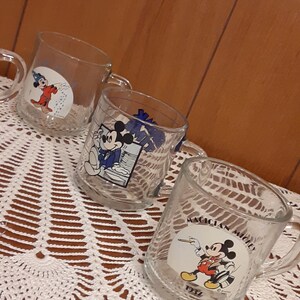 Puede incluir: Tres tazas de vidrio transparente con dise&ntilde;os de Mickey Mouse. Las tazas presentan diferentes im&aacute;genes de Mickey Mouse, incluyendo &eacute;l como mago, director de orquesta y mago. Las tazas est&aacute;n sobre un mantel de ganchillo blanco.