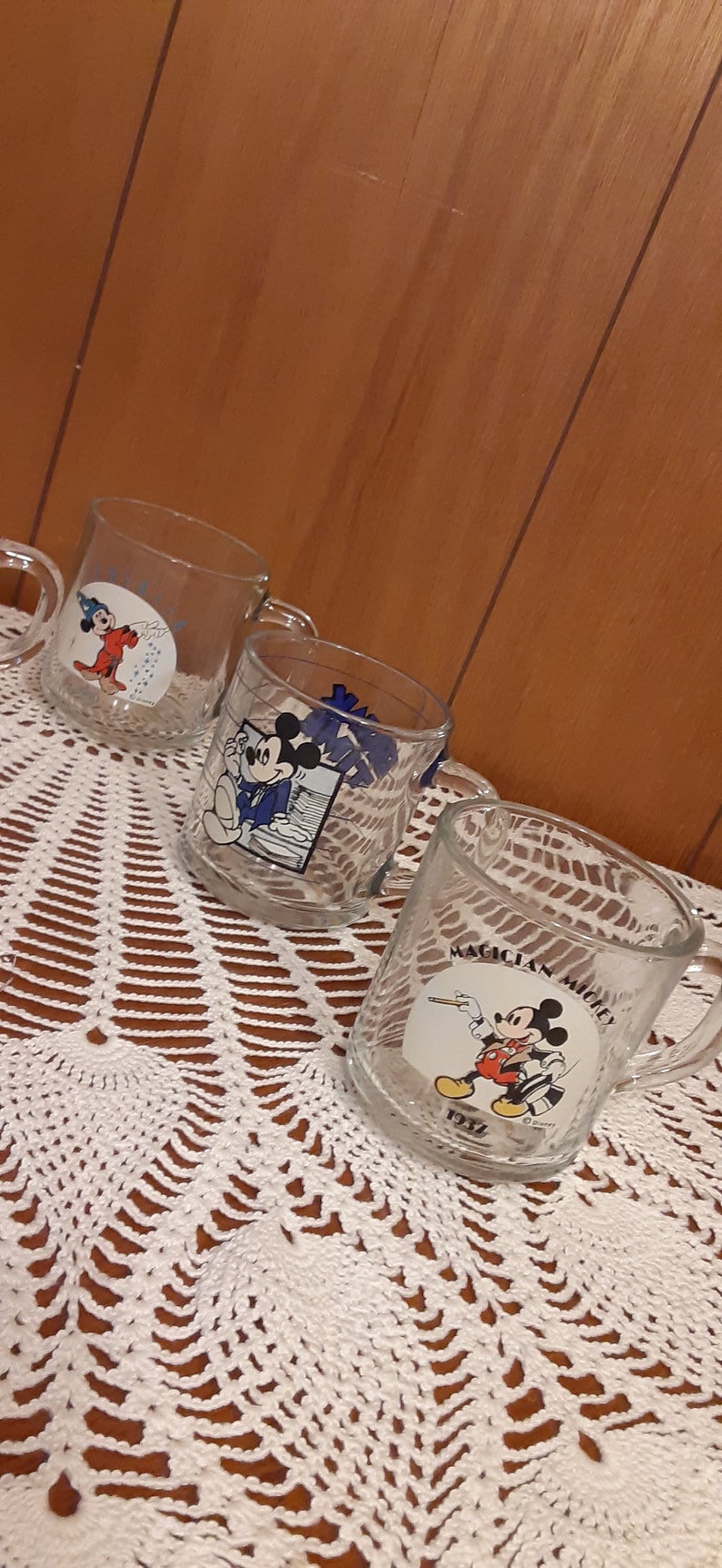 Puede incluir: Tres tazas de vidrio transparente con dise&ntilde;os de Mickey Mouse. Las tazas presentan diferentes im&aacute;genes de Mickey Mouse, incluyendo &eacute;l como mago, director de orquesta y mago. Las tazas est&aacute;n sobre un mantel de ganchillo blanco.