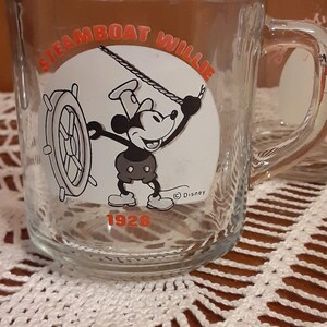 Puede incluir: Una taza de vidrio transparente con una imagen en blanco y negro de Mickey Mouse como Steamboat Willie. La taza tiene el texto "Steamboat Willie" y "1928" en letras naranjas. La taza tambi&eacute;n tiene el s&iacute;mbolo de copyright de Disney.